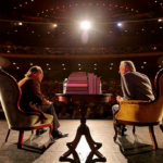 Dr. Lawrence Krauss and Dr. Richard Dawkins sitting together on stage with a spotlight shining down on them.